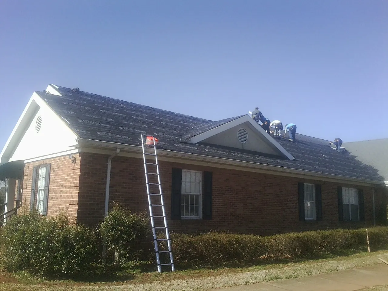 Crew tearing shingles off a brick ranch roof with a ladder set — Metal Roof Repair work by our crew in Serena